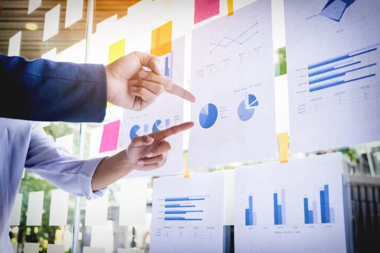 dedos apontando para o gráfico em uma folha pendurada colada em um quadro onde tem mais folhas de gráficos financeiros.