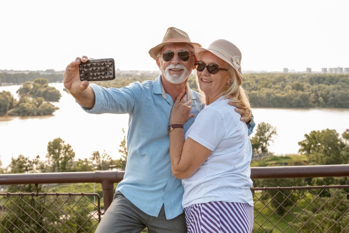 Casal de idosos felizes usando chapéus e óculos de sol, tirando uma selfie com paisagem arborizada e um rio ao fundo. Curtindo sua aposentadoria planejada.